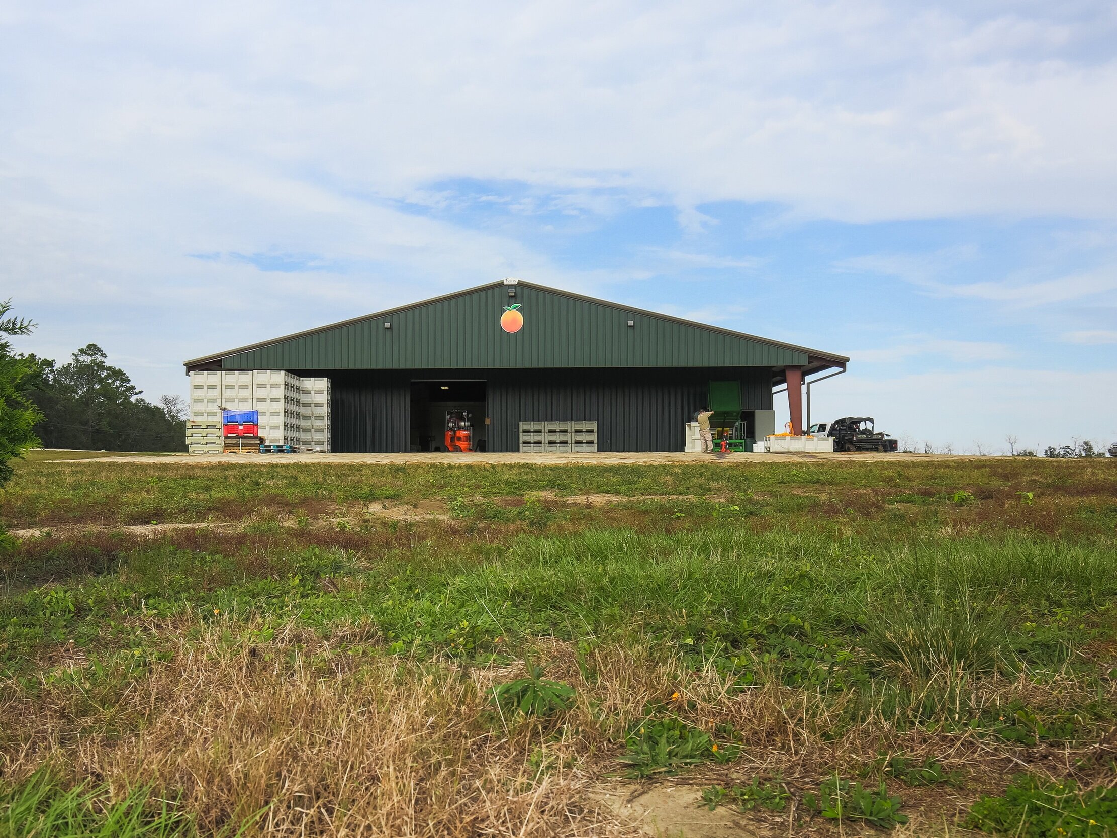 Green agricultural storage facility with crates outside, set against a cloudy sky. The building features a peach logo and is surrounded by grassy terrain, indicating a focus on fruit farming or processing.