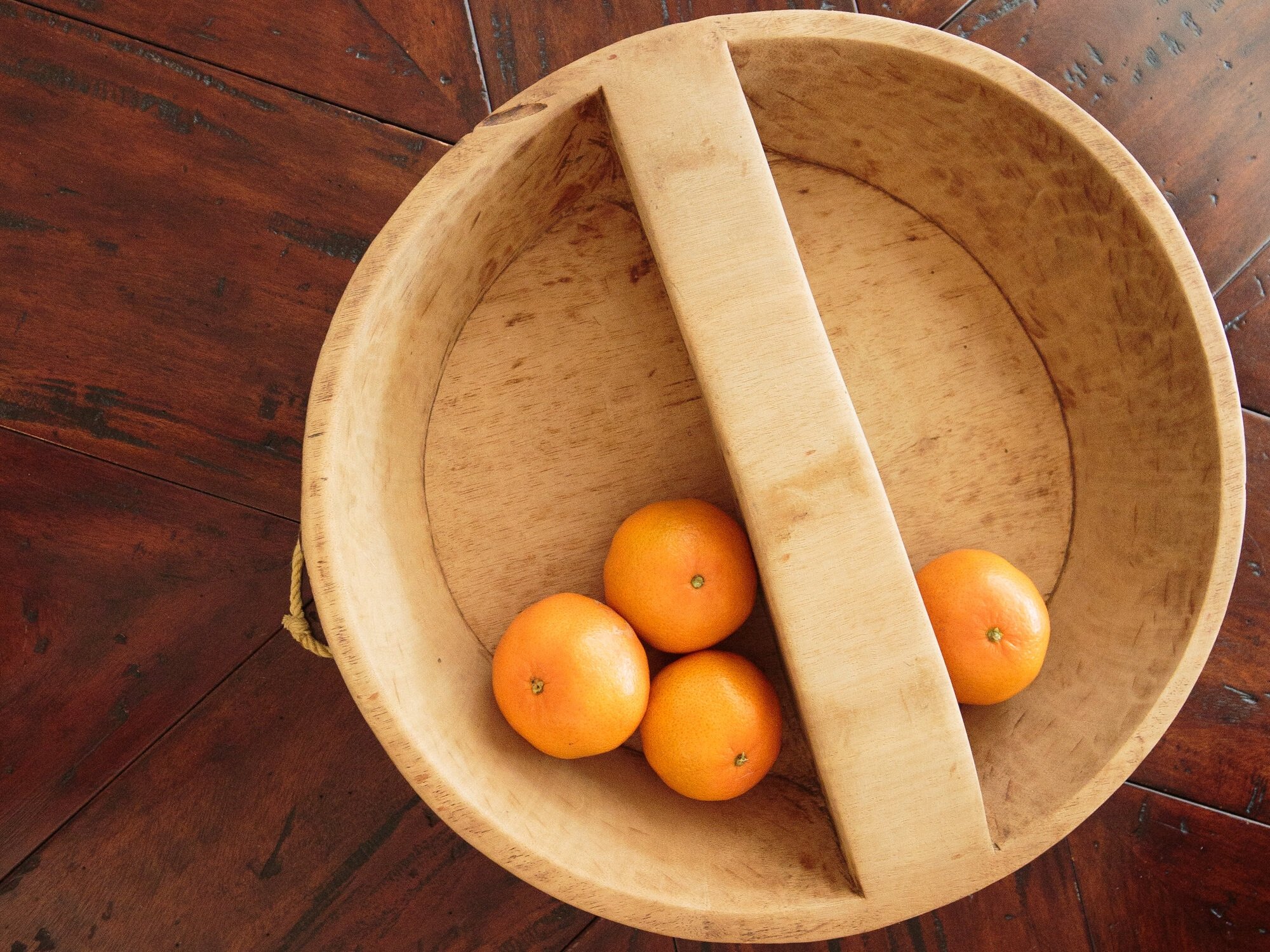 Wooden bowl with a handle containing four fresh oranges, placed on a rustic wooden table.