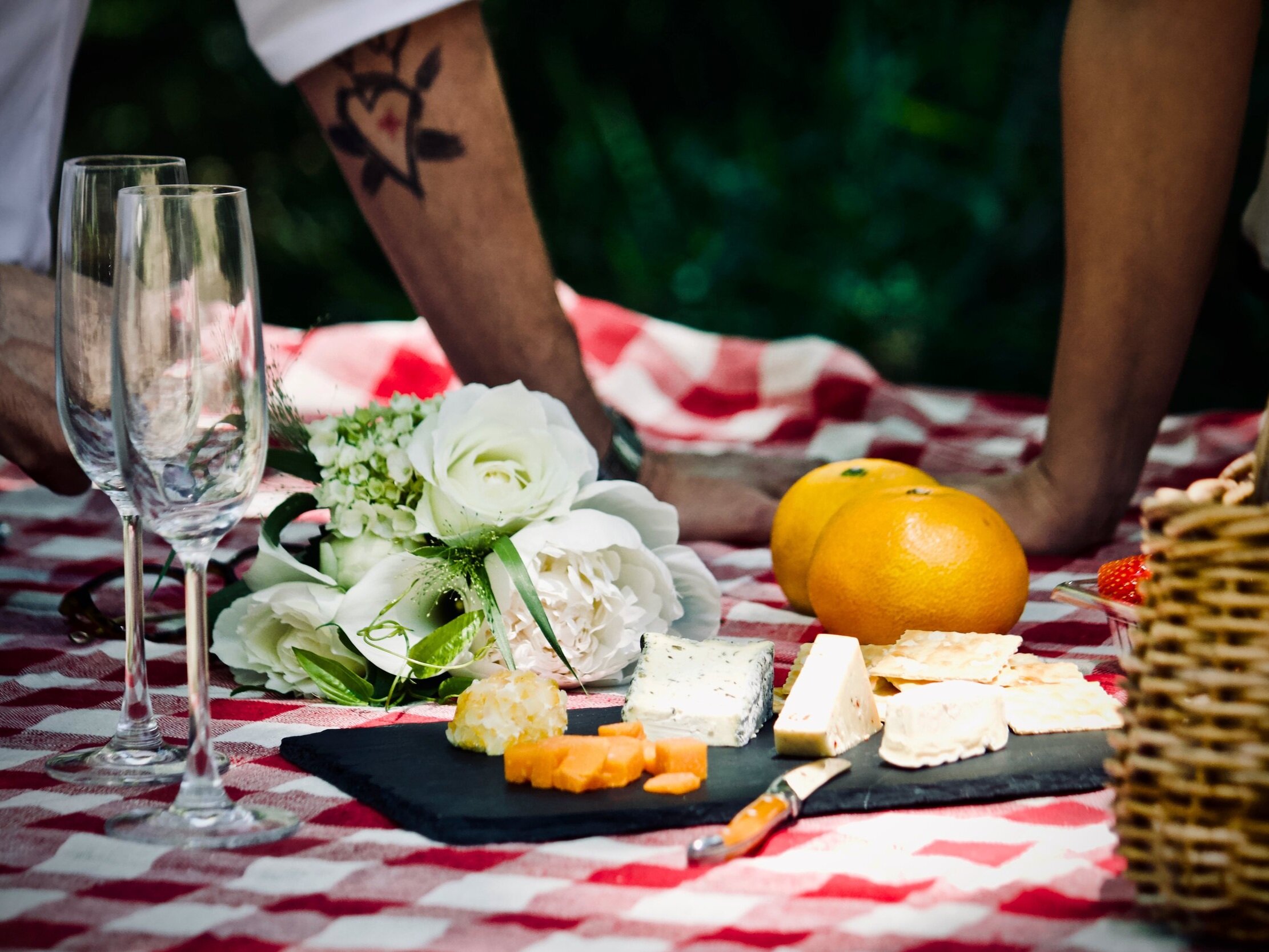 A picnic setup featuring a checkered tablecloth with an assortment of cheeses, crackers, and fresh fruits like oranges and strawberries. Elegant glassware and a bouquet of white roses are placed nearby, creating a romantic outdoor dining atmosphere.